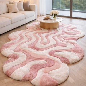 May include: A soft, abstract rug with a wavy pattern in pink and white. The rug is placed on a light wooden floor, creating a cosy feel. The rug's unique shape and colour palette add a modern touch to the room. The rug's dimensions are approximately 200cm x 150cm.