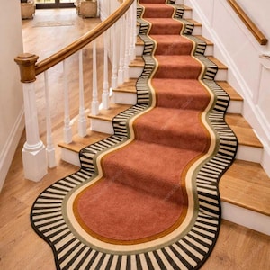 May include: A staircase with a runner rug. The rug is a muted red with a wavy design and a border of black and white stripes, beige, and olive green. The stairs are made of wood and have a wooden handrail.