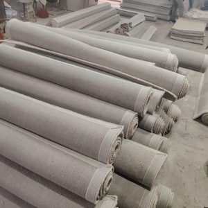 Puede incluir: Una gran colecci&oacute;n de rollos de tela de color gris claro apilados en un almac&eacute;n. Cada rollo presenta una textura suave y uniforme y una forma cil&iacute;ndrica, con los extremos mostrando las capas estrechamente enrolladas del material textil.
