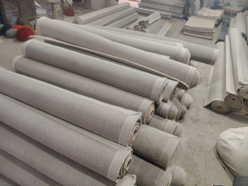 Puede incluir: Una gran colecci&oacute;n de rollos de tela de color gris claro apilados en un almac&eacute;n. Cada rollo presenta una textura suave y uniforme y una forma cil&iacute;ndrica, con los extremos mostrando las capas estrechamente enrolladas del material textil.