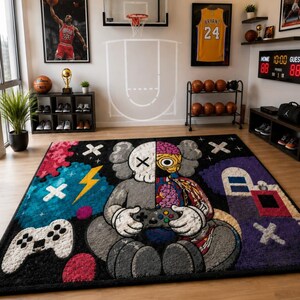 Puede incluir: Una alfombra rectangular que presenta un personaje de dise&ntilde;o dividido sosteniendo un mando de videojuegos. El personaje es mitad gris y mitad anat&oacute;mico con cerebro y tejido muscular expuestos. El fondo incluye un mando blanco, un rayo amarillo y formas abstractas coloridas sobre un campo negro.