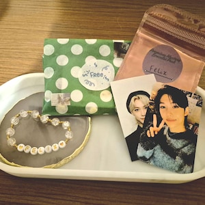 May include: A white oval tray displays a bracelet, wrapped gift, and two photo cards. The bracelet has clear beads and gold letter beads spelling "FELIX". The gift is wrapped in green paper with white polka dots. The photo cards feature two people.