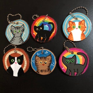 May include: Six wooden cat keychains with different colored backgrounds and painted cat designs. The keychains are round and have a silver chain attached.