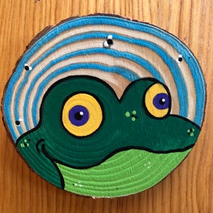 May include: A wooden circle with a blue and white striped background and a green frog painted on top. The frog has yellow eyes with purple pupils and a green body.
