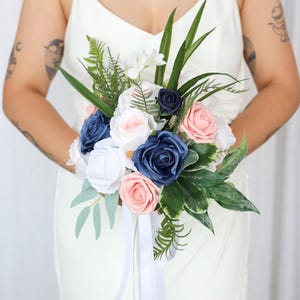 Puede incluir: Un ramo de flores con rosas artificiales en tonos azul marino, rosa suave y blanco, acentuado con frondas de helecho verde y hojas largas y puntiagudas. El arreglo está atado con largas cintas blancas que cuelgan de la base de los tallos.