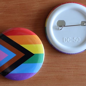 Op de afbeelding: Een ronde knop met een regenboog-pride-vlagontwerp met een zwarte en bruine chevron. De achterkant van de knop is wit met een zilveren pin en de tekst "DC-50".