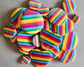 Puede incluir: Un conjunto de botones con rayas de arcoíris en varias formas, incluyendo diseños redondos y en forma de corazón. Los botones presentan una vibrante gama de colores, incluyendo rojo, naranja, amarillo, verde, azul y morado, dispuestos en rayas horizontales.