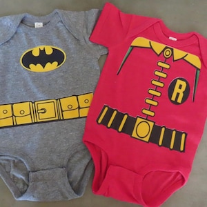 May include: Two short-sleeved baby bodysuits. One is heather gray with a black and yellow Batman logo and a yellow utility belt graphic. The other is red with a yellow collar, a black belt, and a black circle containing the letter R in yellow.