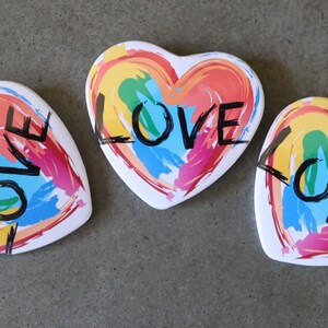 May include: Three heart-shaped ceramic coasters with a rainbow paint design and the word "LOVE" written in black across each coaster.