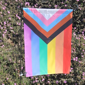 May include: A pride flag with a black chevron on a rainbow background. The flag is attached to a black metal pole.