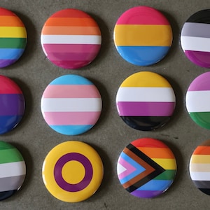 Puede incluir: Doce botones redondos con diferentes banderas de orgullo. Las banderas incluyen las banderas arcoíris, transgénero, bisexual, lesbiana, genderqueer, pansexual, asexual y no binaria.