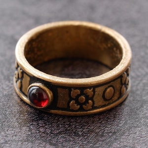 Peut inclure: Une bague en métal doré présentant une bande sombre en creux avec un motif répétitif de petites fleurs et de formes circulaires. Une pierre précieuse unique, ronde, lisse et rouge foncé est sertie sur le devant de la bague, contrastant avec le métal.
