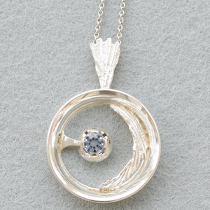 Op de afbeelding: Een zilverkleurige hanger aan een ketting met een cirkelvormig frame met een gestructureerd, organisch ogend detail aan de zijkant. Een enkele, kleine, lichtblauwe ronde edelsteen is in het midden van de cirkel geplaatst, bevestigd door een dunne arm aan de binnenrand.