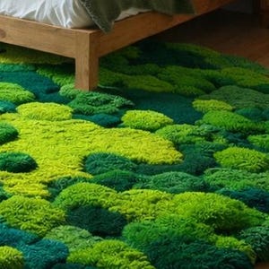 May include: A round rug with a textured, moss-like appearance. The rug features varying shades of green, from light lime to deep teal, creating a natural, organic look. The rug is placed on a wooden floor, partially under a bed.