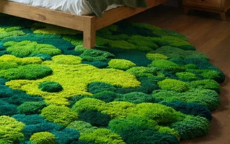 May include: A round rug with a textured, moss-like appearance. The rug features varying shades of green, from light lime to deep teal, creating a natural, organic look. The rug is placed on a wooden floor, partially under a bed.