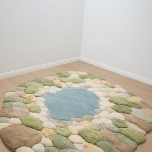 May include: A round rug with a unique design resembling a pond surrounded by stones. The center features a light blue area, with the perimeter composed of various colors of stones, including green, beige, and brown. The rug is placed on a wooden floor.