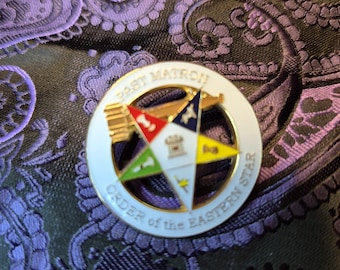 May include: A white and gold Order of the Eastern Star pin with the words "Past Matron" and "Order of the Eastern Star" encircling a colorful star emblem. The pin is set against a paisley patterned background.