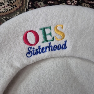 Puede incluir: Boina blanca con el texto bordado "OES Sisterhood" en rojo, verde, amarillo y azul. La boina está hecha de un material suave y difuso.