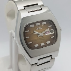 May include: A vintage silver-toned Jemis automatic watch. The watch face is brown with silver hands and markers. The day and date are displayed at the 3 o'clock position. The watch has a stainless steel band.