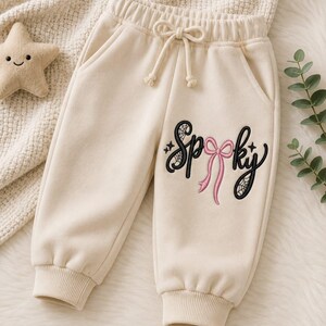 May include: Cream-colored sweatpants featuring an elastic waistband with a drawstring and ribbed cuffs. The left leg displays black embroidered text that reads Spooky, with a pink bow replacing the letter o and spiderweb details.