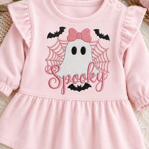 May include: A light pink long-sleeve dress with ruffled shoulders and a round neckline featuring two snap buttons. The center displays an embroidered white ghost with a pink bow, black bats, a spider web, and the word Spooky in pink script.
