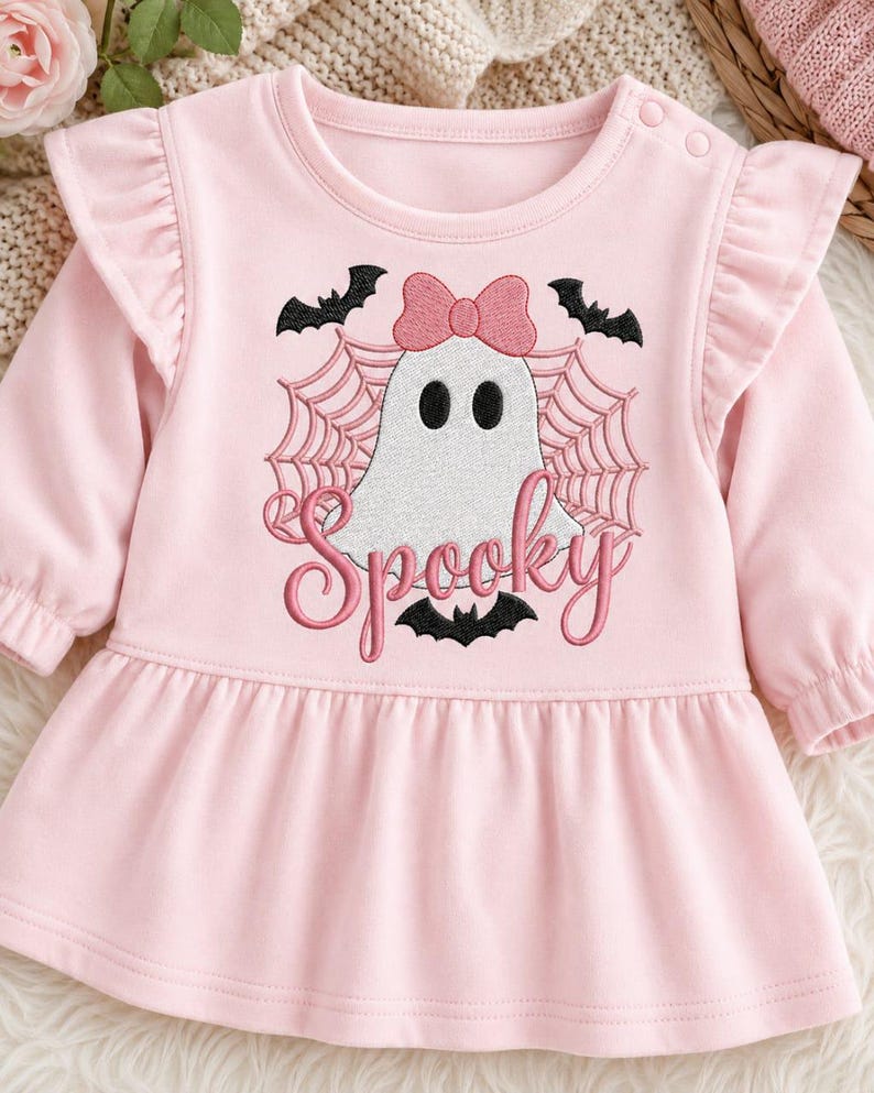 May include: A light pink long-sleeve dress with ruffled shoulders and a round neckline featuring two snap buttons. The center displays an embroidered white ghost with a pink bow, black bats, a spider web, and the word Spooky in pink script.