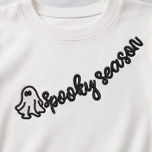 May include: An off-white crewneck sweatshirt featuring black embroidery on the chest. The design includes a small ghost figure positioned to the left of the text "Spooky season" written in a cursive, black embroidered font across the front.