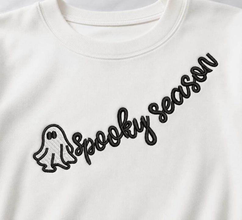 May include: An off-white crewneck sweatshirt featuring black embroidery on the chest. The design includes a small ghost figure positioned to the left of the text "Spooky season" written in a cursive, black embroidered font across the front.