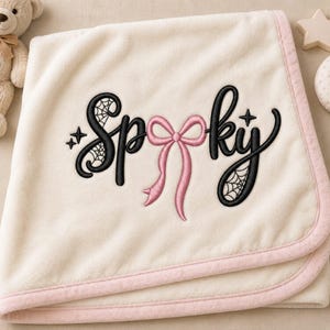 May include: A cream-colored fleece blanket with a light pink trim. The center features black embroidered text that reads Spooky, with a pink bow replacing the letter o and spiderweb patterns integrated into the letters S and y, plus small stars.