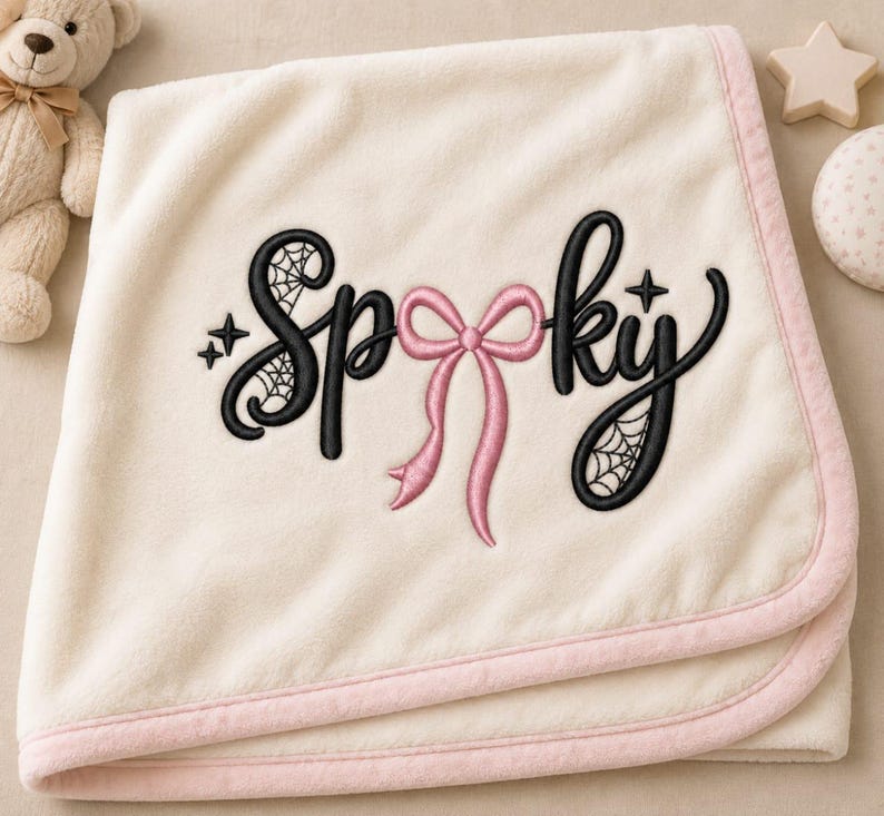 May include: A cream-colored fleece blanket with a light pink trim. The center features black embroidered text that reads Spooky, with a pink bow replacing the letter o and spiderweb patterns integrated into the letters S and y, plus small stars.