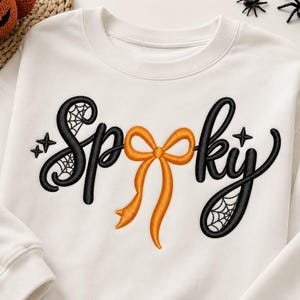 May include: A white crewneck sweatshirt featuring black embroidered text that reads Spooky. The letter O is replaced by an orange ribbon bow. The letters S and Y include black spiderweb patterns and small star accents within the embroidery.