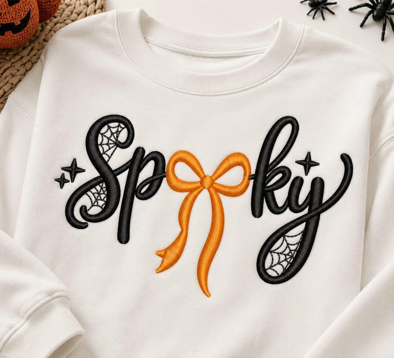 May include: A white crewneck sweatshirt featuring black embroidered text that reads Spooky. The letter O is replaced by an orange ribbon bow. The letters S and Y include black spiderweb patterns and small star accents within the embroidery.