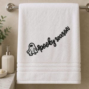 May include: A white bath towel featuring black embroidery of a small ghost and the text "spooky season" written in a cursive script. The towel has a textured border with three horizontal lines near the bottom edge and hangs on a metal bar.