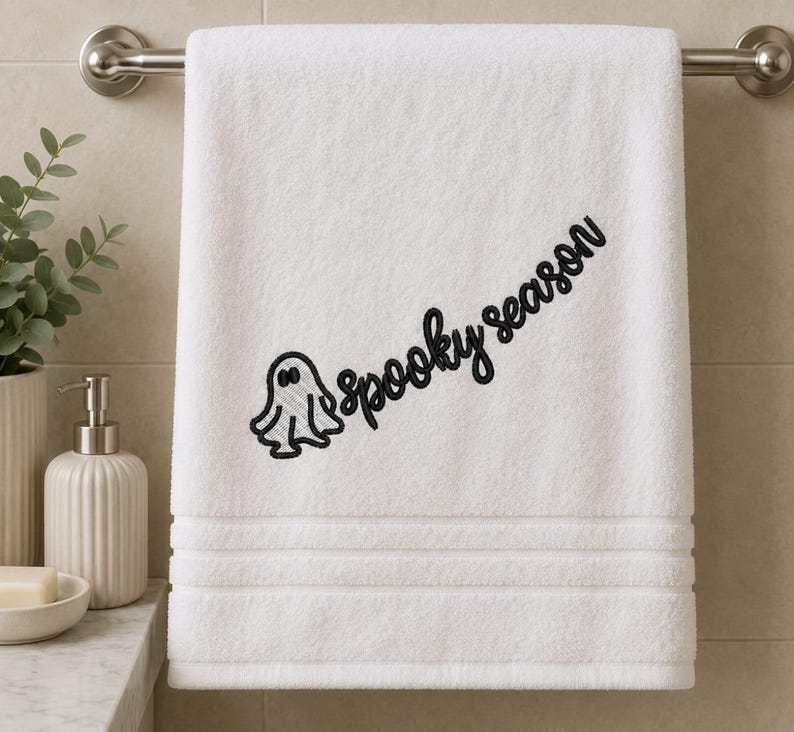 May include: A white bath towel featuring black embroidery of a small ghost and the text "spooky season" written in a cursive script. The towel has a textured border with three horizontal lines near the bottom edge and hangs on a metal bar.