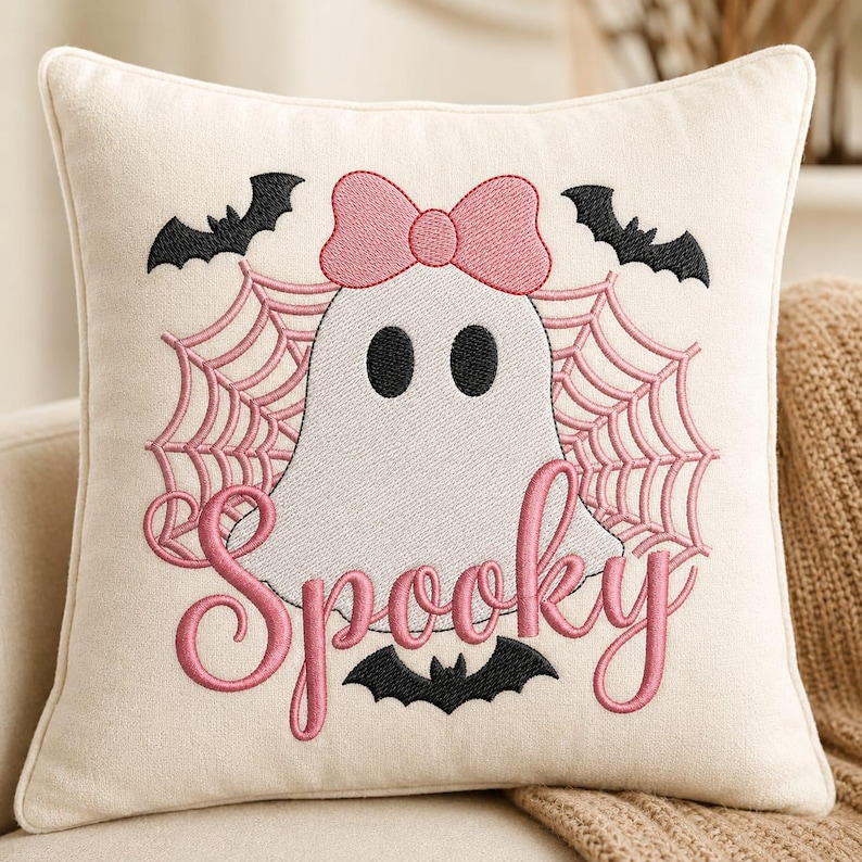 May include: A square off-white throw pillow featuring an embroidered design of a white ghost with a pink bow, surrounded by pink spiderwebs and black bats. The word Spooky is embroidered in pink cursive script below the ghost figure.