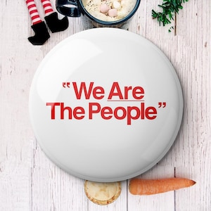 May include: A white button with the red text "We Are The People". The button is on a light wood surface, with a carrot, a small pie, and a mug of hot chocolate with marshmallows. Festive holiday theme.