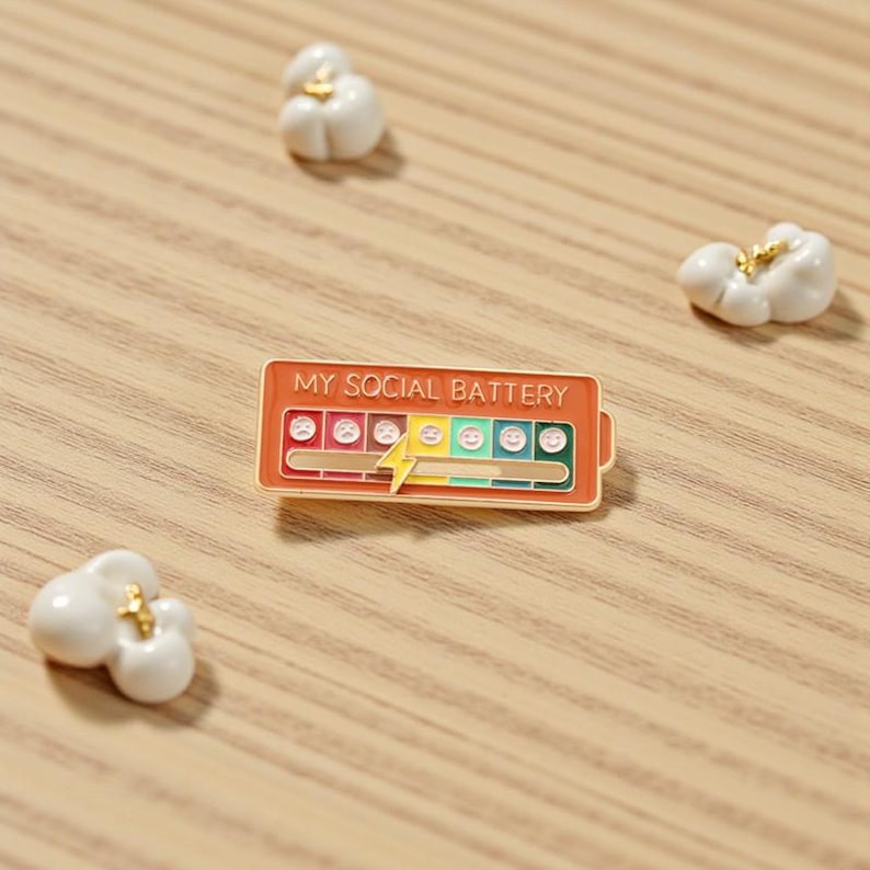 May include: An enamel pin with the text "MY SOCIAL BATTERY" in orange, featuring a battery level indicator with emoji faces. The pin has a gold border and a lightning bolt design. Scattered around the pin are three small, white popcorn kernels with gold accents.