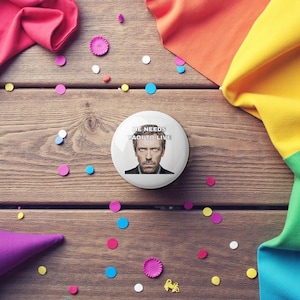 Puede incluir: Un botón blanco con el texto "HE NEEDS YAOI TO LIVE" y una foto de un hombre. El botón está rodeado de confeti colorido y tela sobre una superficie de madera.