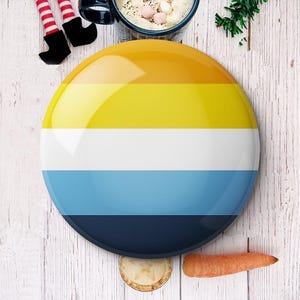 May include: A large, round button with a glossy finish, featuring horizontal stripes in shades of yellow, white, blue, and navy. The button is placed on a light wood-grain surface, with a carrot, pie, and a mug of hot chocolate nearby.