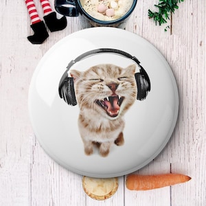 以下が含まれることがあります： 黒いヘッドホンをつけたトラ猫が、喜びの表情で口を大きく開けている白いボタン。猫はボタンの中央に配置され、白い背景があります。ニンジンと小さなパイが前景にあります。