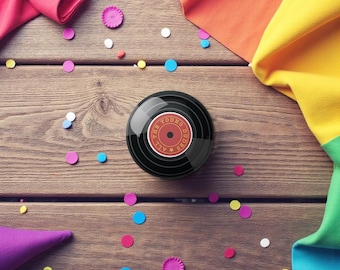 May include: A black, round object resembling a vinyl record with the text "THE YOUNG DUDES ALL" in red. The object is surrounded by colorful confetti and fabric on a wooden surface.