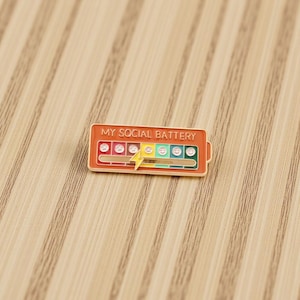 Puede incluir: Pin rectangular de esmalte con el texto "MY SOCIAL BATTERY". El pin presenta un indicador de nivel de batería con emoticonos y un rayo. El pin es de color naranja con un borde dorado.