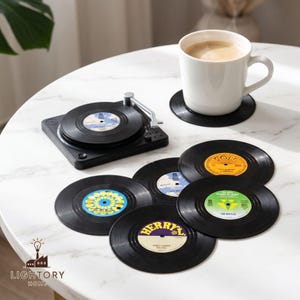 Puede incluir: Un juego de posavasos de vinilo negro con etiquetas centrales coloridas, incluyendo uno colocado bajo una taza de cerámica blanca y otro sobre un soporte negro con forma de tocadiscos. Las etiquetas tienen diseños.