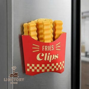 Könnte beinhalten: Ein neuartiger Kühlschrankmagnet in Form einer Pommes-Frites-Schachtel. Der rote Karton trägt die Worte "FRIES Clips" in einer Retro-Schrift, mit gelben Pommes, die herausquellen. Der Magnet ist an einem Edelstahlkühlschrank befestigt.