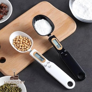Puede incluir: Dos básculas de cocina digitales con cabezales en forma de cuchara. Una es blanca, mostrando 19.6, la otra negra, mostrando 6.6. Ambas tienen pantallas LCD y botones. Una cuchara contiene frijoles, la otra polvo blanco. Medidas en gramos.
