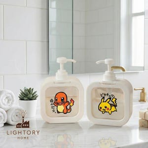 May include: Two off-white, square-shaped soap dispensers with white pump tops. One features a Charmander graphic and Japanese text, the other a Pikachu graphic and Japanese text. The dispensers are on a white countertop.
