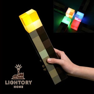 Op de afbeelding: Een LED-fakkel lamp in Minecraft-stijl met een geel verlichte bovenkant en een kleurblokontwerp op de handgreep. De lamp wordt door een hand vastgehouden. Extra kubusvormige lampen in verschillende kleuren zijn op de achtergrond te zien. Het logo "LIGHTORY HOME" staat onderaan.