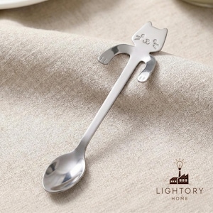 Puede incluir: Una cuchara de acero inoxidable con un diseño en forma de gato en el mango. El gato tiene una cara sonriente y brazos y piernas extendidos. La cuchara está sobre una superficie de lino beige. Utensilio de cocina.