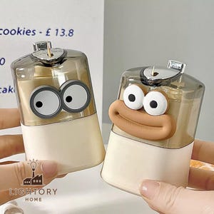 Puede incluir: Dos dispensadores de palillos de dientes novedosos con caras de dibujos animados. Los dispensadores tienen una parte superior transparente, una base beige y una parte superior de metal plateado. Uno tiene ojos saltones grandes y el otro una cara sonriente con una boca grande.
