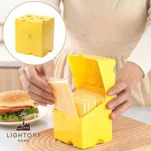 Puede incluir: Un cortador de queso amarillo con forma de bloque de queso suizo, con agujeros en la parte superior y una tapa con bisagras. El cortador está abierto, revelando rebanadas de queso envueltas individualmente. Una hamburguesa está en el fondo.